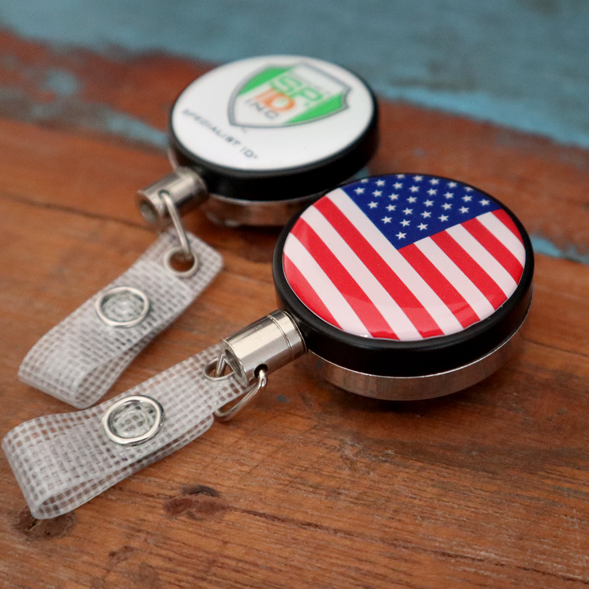 Two Heavy Duty Custom Printed Badge Reels With Steel Cable 2120-3305 - Add Your Logo lie on a wooden surface. One features an American flag design, while the other sports a green and white logo with text, promoting brand awareness. Both have transparent clips attached for easy use.
