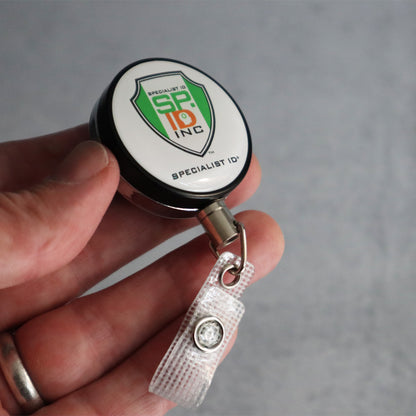Close-up of a hand holding a Heavy Duty Custom Printed Badge Reel With Steel Cable 2120-3305 - Add Your Logo with a clip and a visible "Specialist ID" logo, promoting brand awareness.