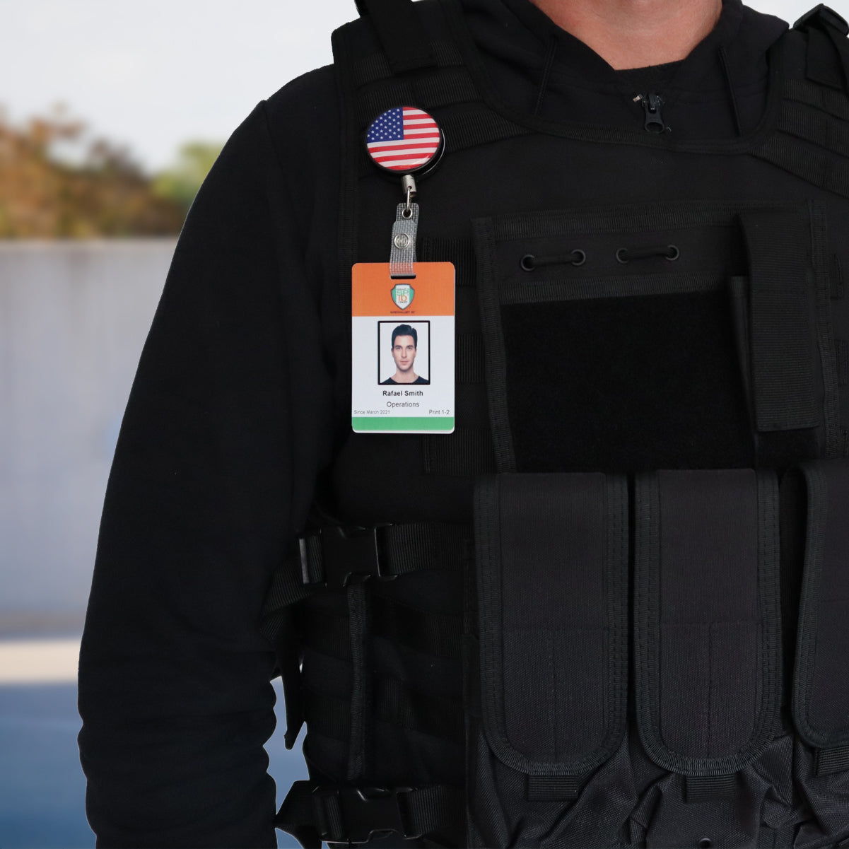 A person wearing a tactical vest with an ID badge attached, featuring a Heavy Duty Custom Printed Badge Reel With Steel Cable 2120-3305 - Add Your Logo and an American flag pin above it. The badge displays a photo and the name "Robert Smith, Operations.