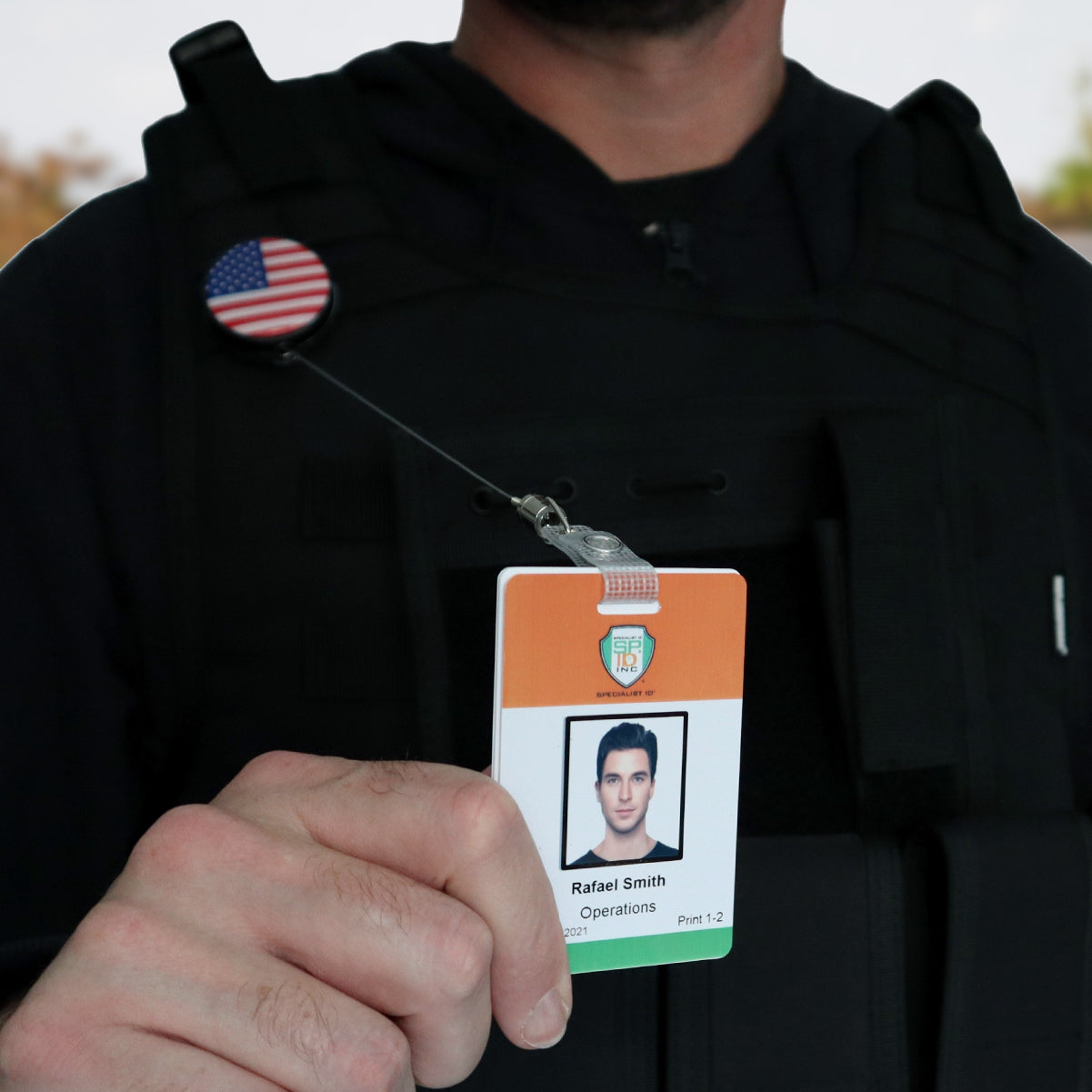 A person wearing a black vest holds an identification card, attached to a Heavy Duty Custom Printed Badge Reel With Steel Cable 2120-3305 - Add Your Logo, showing a photo of "Rafael Smith" with the text "Operations" on it. The vest also has a round pin with the American flag.