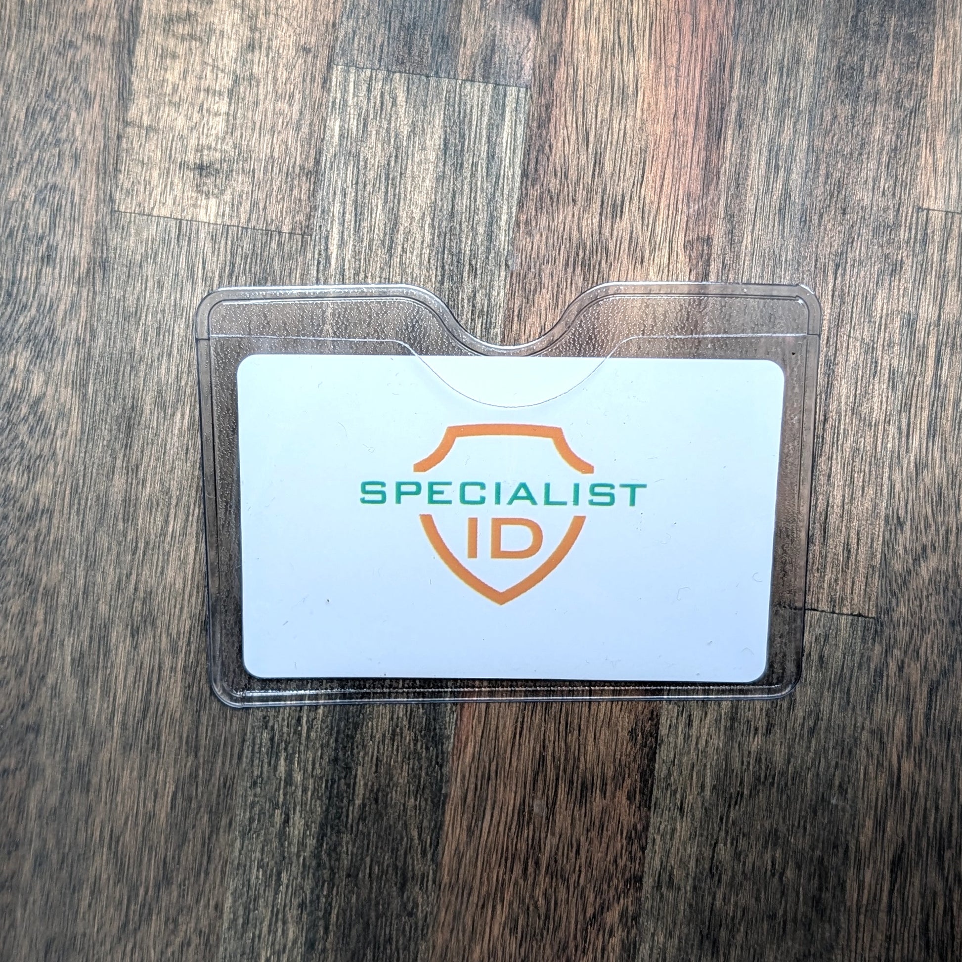 A Horizontal Textured Vinyl Badge Holder with Clip (P/N 1810-1000) secures a “SPECIALIST ID” card with a shield logo, all shown on a wooden surface.