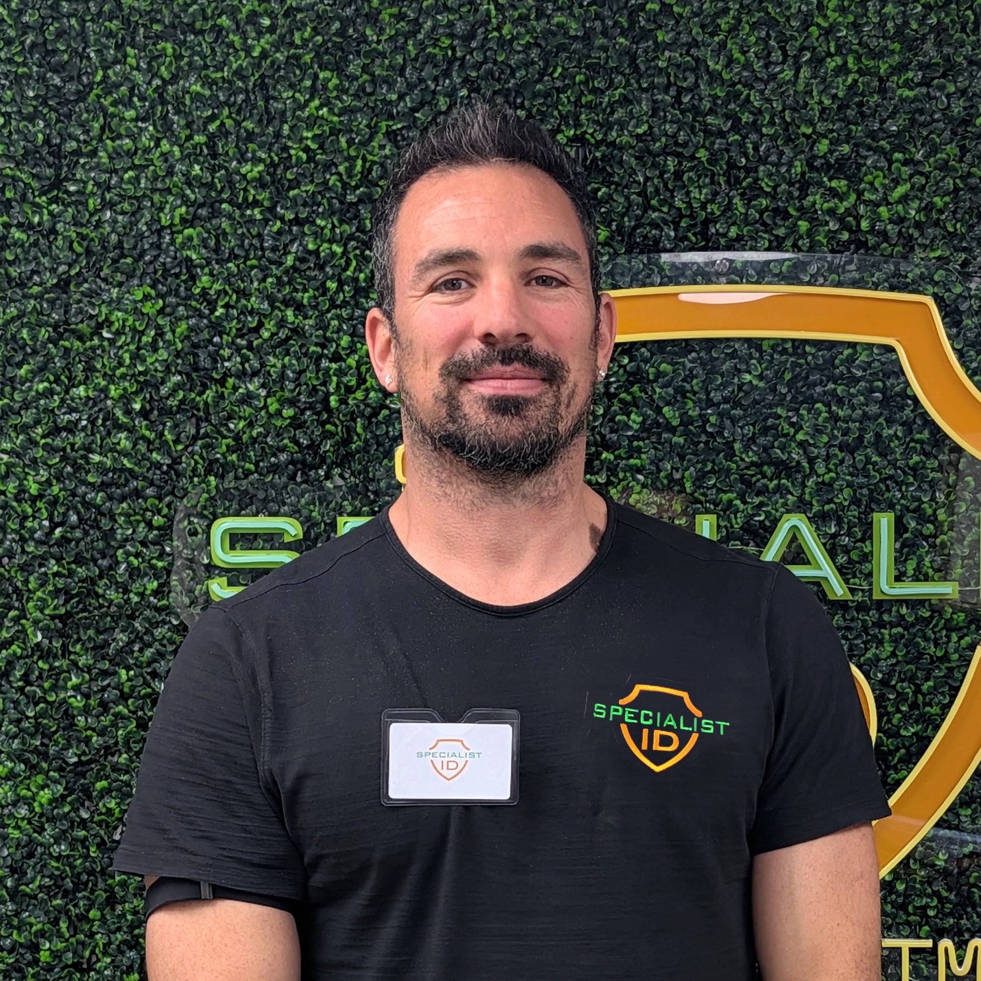 A man with short dark hair and a beard stands in front of a green wall with a "Specialist ID" logo, wearing a black "Specialist ID" shirt and a name badge secured in the Horizontal Textured Vinyl Badge Holder with Clip (P/N 1810-1000).