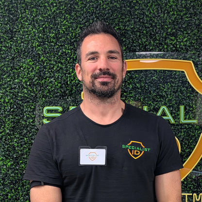 A man with short dark hair and a beard stands in front of a green wall with a "Specialist ID" logo, wearing a black "Specialist ID" shirt and a name badge secured in the Horizontal Textured Vinyl Badge Holder with Clip (P/N 1810-1000).