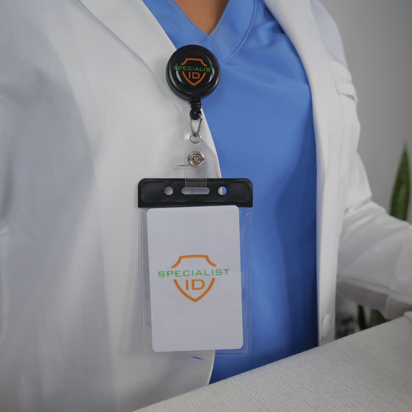 A person in a blue top and white coat wears a name badge with a retractable holder. The badge is displayed in the Vertical Vinyl Badge Holder with Color Bar, Standard ID Badge Size (1820-105X), showing the text "SPECIALIST ID" and a shield logo.