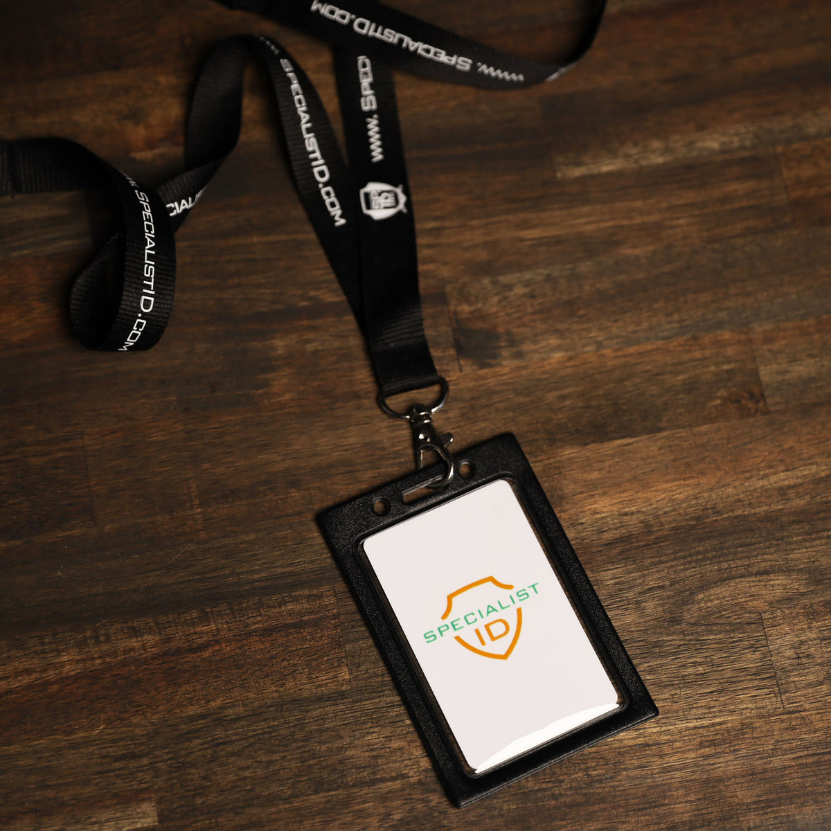 Black lanyard with “SPECIALISTID.COM” text attached to a black Vertical Vinyl Color Frame Badge Holder (P/N 1820-300x) displaying a card labeled “SPECIALIST ID” on a wooden surface—perfect for use with color-coded access systems.