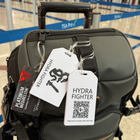 A black travel bag with a "HYDRA FIGHTER" tag and a Custom Rigid Plastic Luggage Tag Holder, personalized with your logo, is displayed at an airport with TSA PreCheck signs, boosting brand awareness.