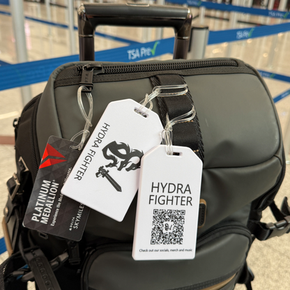 A black travel bag with a "HYDRA FIGHTER" tag and a Custom Rigid Plastic Luggage Tag Holder, personalized with your logo, is displayed at an airport with TSA PreCheck signs, boosting brand awareness.