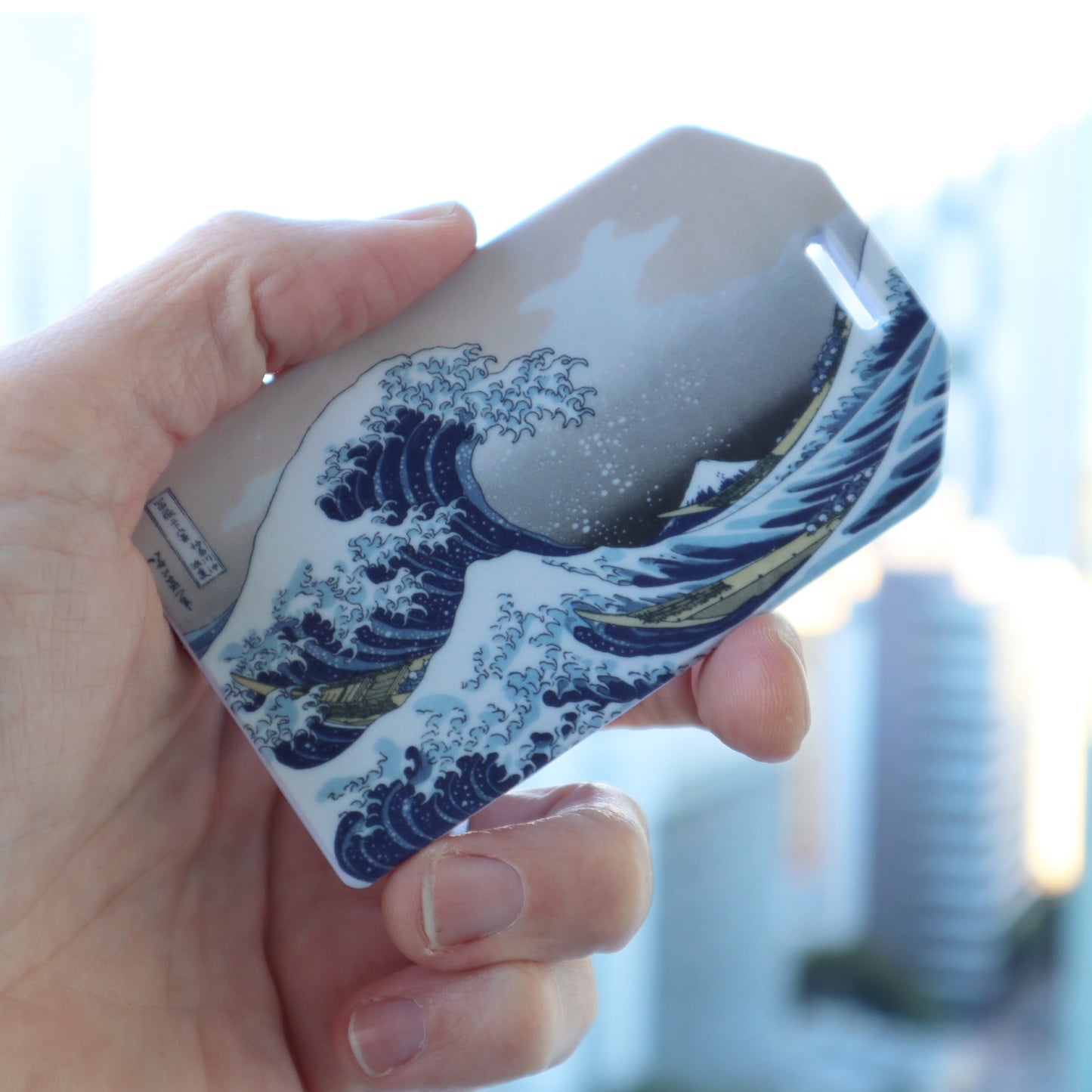 A hand holds a rectangle-shaped luggage tag resembling the Katsushika Hokusai-inspired "Great Wave" design, complete with a clear loop,