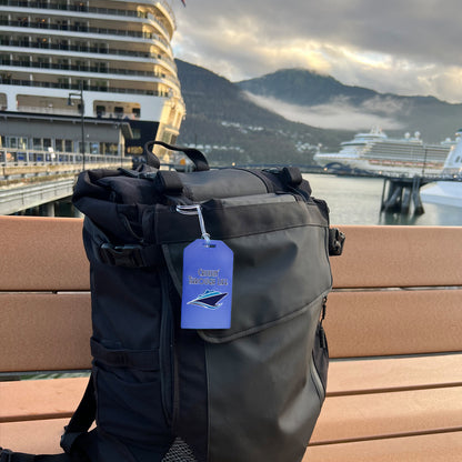 A black backpack, featuring the "Cruisin' Through Life" Hard Plastic Luggage Tag with a cruise ship design, rests on a bench against the majestic backdrop of a cruise ship. It's the ideal travel accessory for those who embrace adventure and the allure of wanderlust at sea.