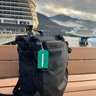 A black backpack with Custom Rigid Plastic Luggage Tag Holders with 6" Clear Loop - Personalize with Your Logo rests on a bench, promoting brand awareness against the backdrop of moored cruise ships and a mountainous landscape.
