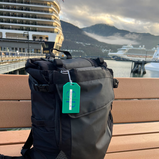 A black backpack with Custom Rigid Plastic Luggage Tag Holders with 6" Clear Loop - Personalize with Your Logo rests on a bench, promoting brand awareness against the backdrop of moored cruise ships and a mountainous landscape.