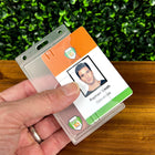 A person is holding an ID card labeled "Raphael Smith, Operations" over a Custom Vertical Rigid Plastic Card Holder (1840-6500) - Personalize with Your Logo, with a green leafy background.
