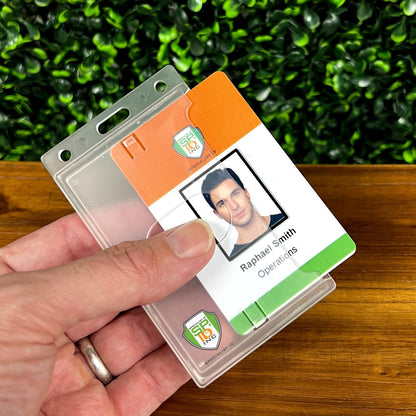A person is holding an ID card labeled "Raphael Smith, Operations" over a Custom Vertical Rigid Plastic Card Holder (1840-6500) - Personalize with Your Logo, with a green leafy background.