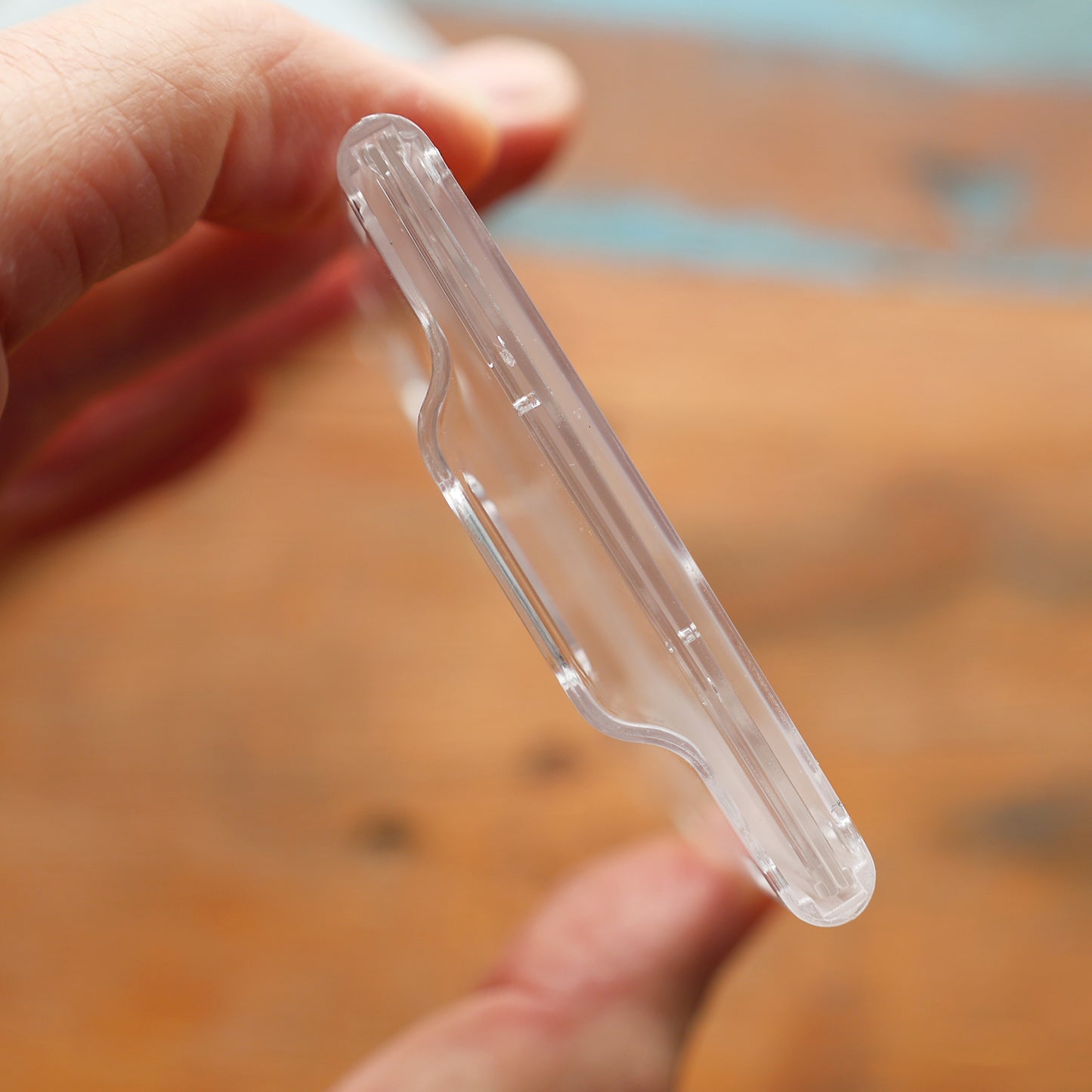 A close-up of a hand holding the sleek Rigidwear Clear Vertical 2-Card Badge Holder 1840-6560 (Jam P/N 706-NN2), featuring a slight indentation in the middle, set against a blurred wooden background.