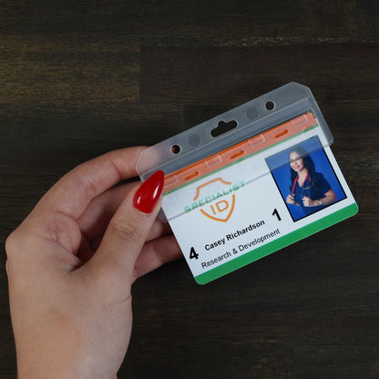 A hand holds an ID badge featuring a photo and the text “Casey Richardson, Research & Development, 4, 1,” secured in a Swipe Card Holder - Rigid Horizontal Half Card Badge Holder - Leaves Mag Stripe Exposed for Easy Swiping (1840-8000), resting on a dark wood surface.