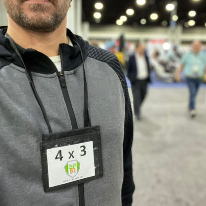 A person wearing a grey and black jacket with a Premium Color 3 Pocket 4x3 Nylon Credential Neck Lanyard Wallet with Adjustable Lanyard (1860-260X) at a convention or event, with other attendees in the background.