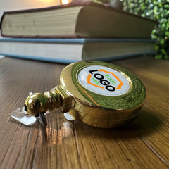 A Silver or Gold Custom Printed Retractable Badge Reel With Belt Clip - Upload Your Logo, perfect for brand awareness, rests on a wooden surface with two stacked books and a plant in the background.