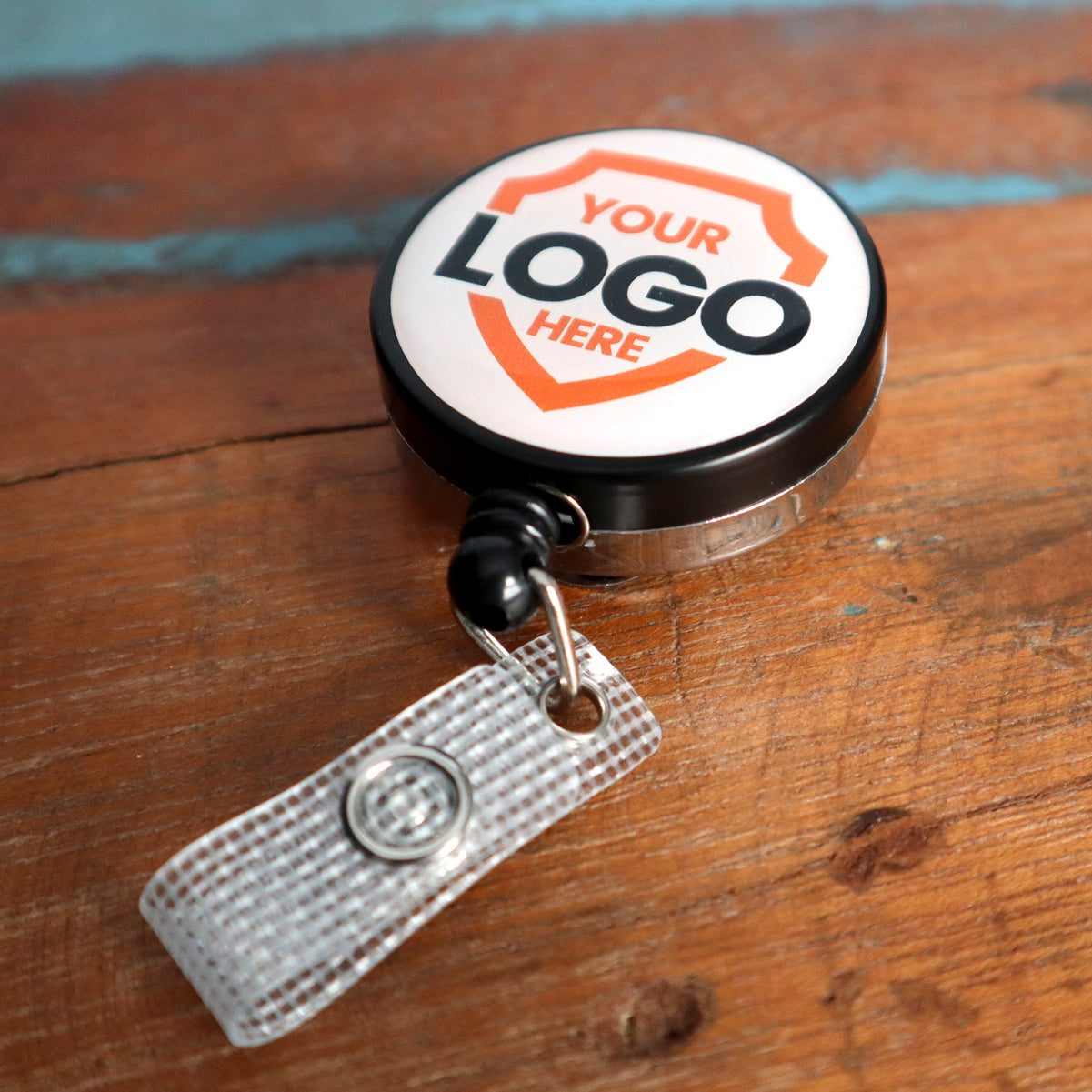 A Custom Badge Reels - Heavy Duty Custom Printed Badge Reel with Belt Clip - Upload Your Logo on a wooden surface features a customizable round top that says "Your Logo Here," perfect for custom badge reels to enhance brand visibility.