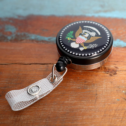 A round black retractable badge holder with a U.S. presidential eagle design, attached to a white clip, rests on a wooden surface—perfect for Custom Badge Reels - Heavy Duty Custom Printed Badge Reel with Belt Clip - Upload Your Logo or promotional giveaways that enhance brand visibility.