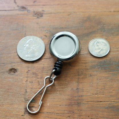 The Mini Chrome Silver Key Reel with Spring Clip and Metal Swivel Hook (2120-3400) is elegantly displayed on a wooden surface, accompanied by a quarter and a nickel.