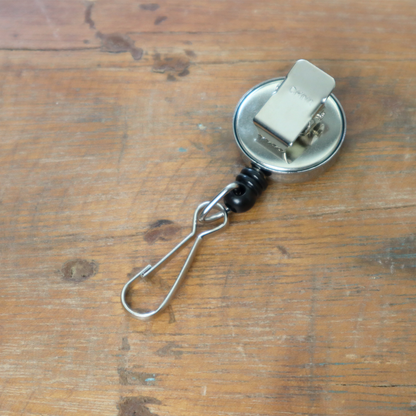 The Mini Chrome Silver Key Reel with Spring Clip and Metal Swivel Hook (2120-3400) is resting elegantly on a wooden surface, its retractable cord perfectly displayed.