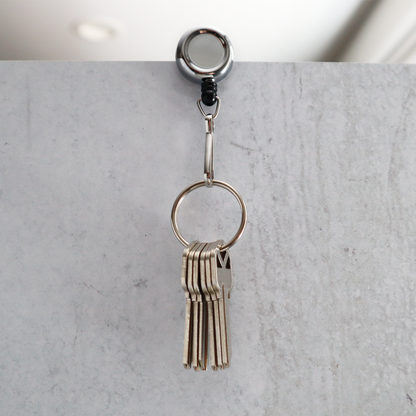 A set of keys dangles from a Mini Chrome Silver Key Reel With Spring Clip And Metal Swivel Hook (2120-3400), its metal swivel hook securely attached to a gray marble surface.