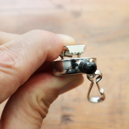 A hand holds a shiny, chrome silver mini key reel with a spring clip and metal swivel hook. The background features a blurred wooden surface, creating an elegant simplicity. Sold by Specialist ID.