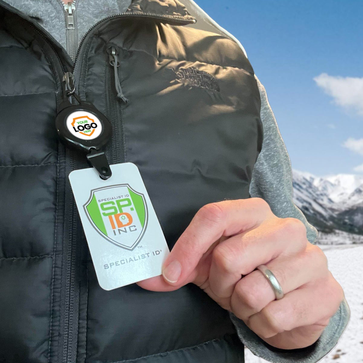 A person wearing a gray zip-up jacket and black vest holds an ID badge lanyard with the "Specialist ID, Inc" logo. A Customi Ski Pass and Sports Retractable Reel - Add Your Logo (2120-4001-Custom) dangles from the lanyard, perfect for quick access. Snow-covered mountains and a blue sky are visible in the background.