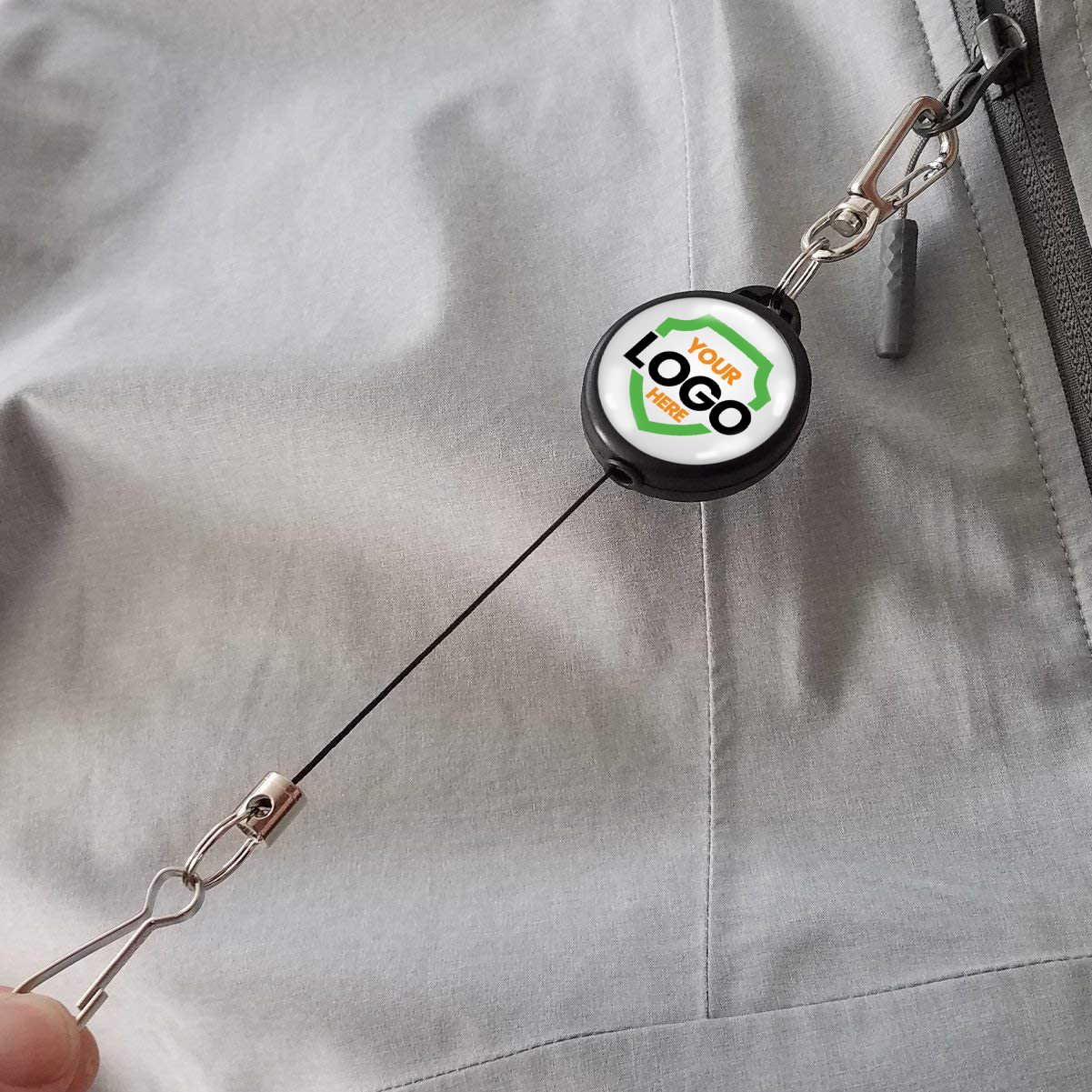A Custom Retractable Ski Pass Holder - Heavy Duty Sport Reel (2120-4101) - Add Your Logo attached to a zipper on a gray garment, featuring a custom logo on its round casing. Sold by Specialist ID.
