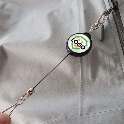 A Custom Retractable Ski Pass Holder - Heavy Duty Sport Reel (2120-4101) - Add Your Logo attached to a zipper on a gray garment, featuring a custom logo on its round casing. Sold by Specialist ID.
