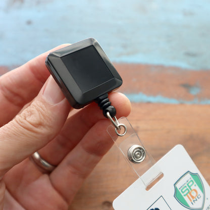 A hand holds the stylish Black Square Badge Reel With Spring Clip (P/N 2120-5701), featuring a retractable nylon cord and a spring clip securely attached to a card.