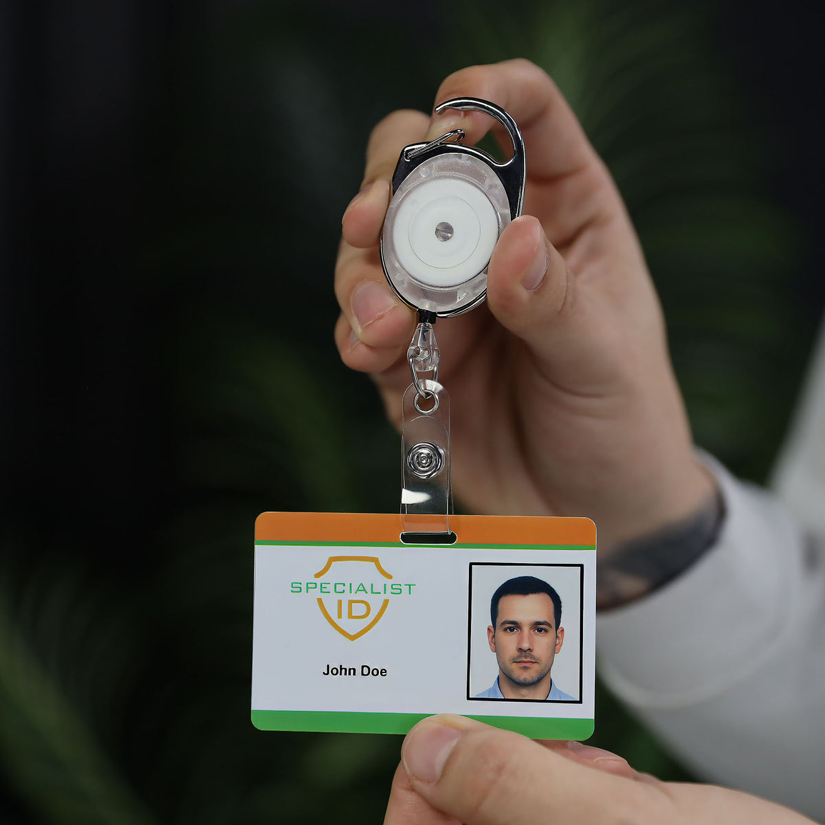 A person holds up a Carabiner Badge Reel with Oval Clip for Belt Loops and Bag Straps (2120-70XX) displaying an ID card labeled “Specialist ID” with a photo and the name “John Doe.”.