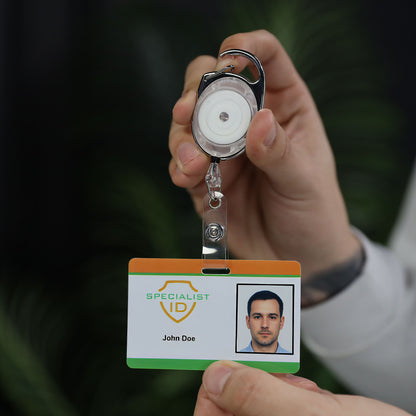 A person holds up a Carabiner Badge Reel with Oval Clip for Belt Loops and Bag Straps (2120-70XX) displaying an ID card labeled “Specialist ID” with a photo and the name “John Doe.”.