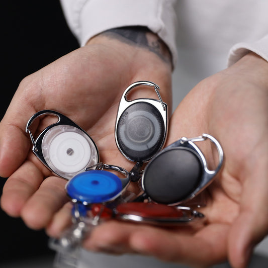 A person holds several Carabiner Badge Reel with Oval Clip for Belt Loops and Bag Straps (2120-70XX) in various colors and designs in their open hands.