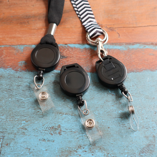 Three Retractable Badge Reels with Top Slot Lanyard Attachment (P/N 2120-7501), featuring a sleek black design and practical vinyl strap clips for extended reach, are lying on a wooden surface.