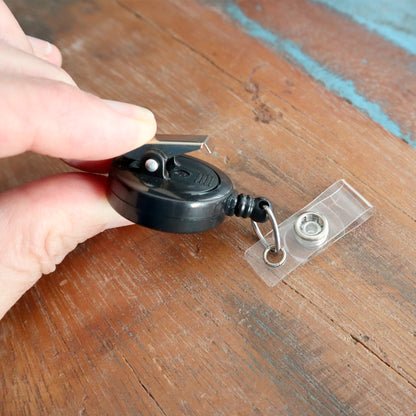 A hand holds the modern Badge Reel with Swivel Spring Clip (P/N 2120-760X), elegantly positioned on a wooden surface. Sold by Specialist ID.