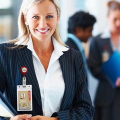 A smiling woman in a pinstripe suit wears Custom Badge Reels with Swivel Spring Clip - Personalized Bulk ID Badge Holders for Business, Nurses, and Teachers and a visible ID badge while standing in a professional setting, with two other people blurred in the background.