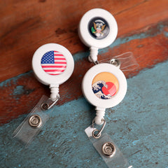 Three Custom Badge Reels with Swivel Spring Clip - Personalized Bulk ID Badge Holders for Business, Nurses, and Teachers with white casings, each featuring a different design: the US flag, a stylized wave and sun, and a presidential seal. Perfect for promotional giveaways, these custom badge reels are clipped onto a weathered wooden surface.