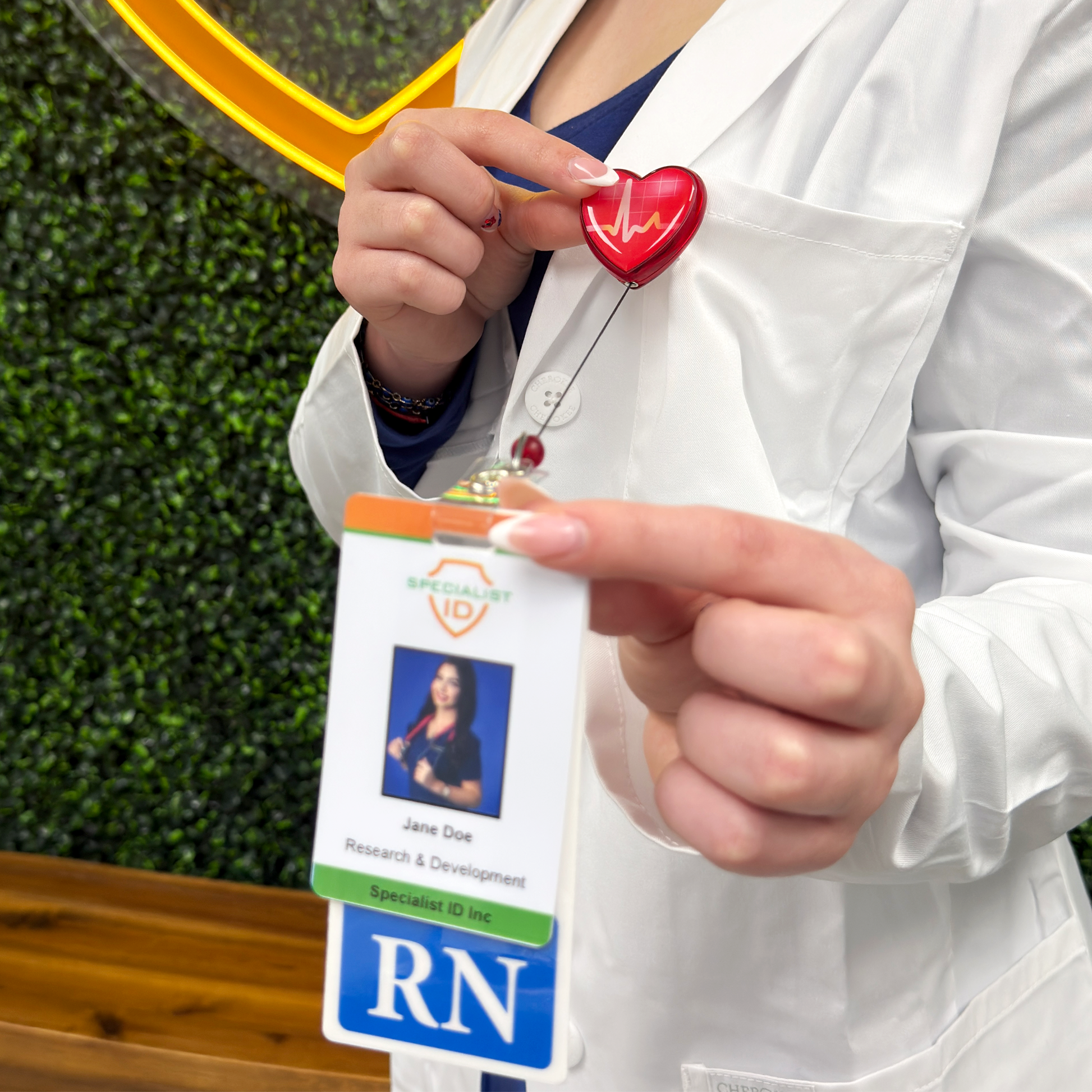 Person in a white coat wearing a red heart-shaped badge reel with heartbeat design and an RN badge extended to show her vertical RN badge buddy with ID badge. Sold by Specialist ID.