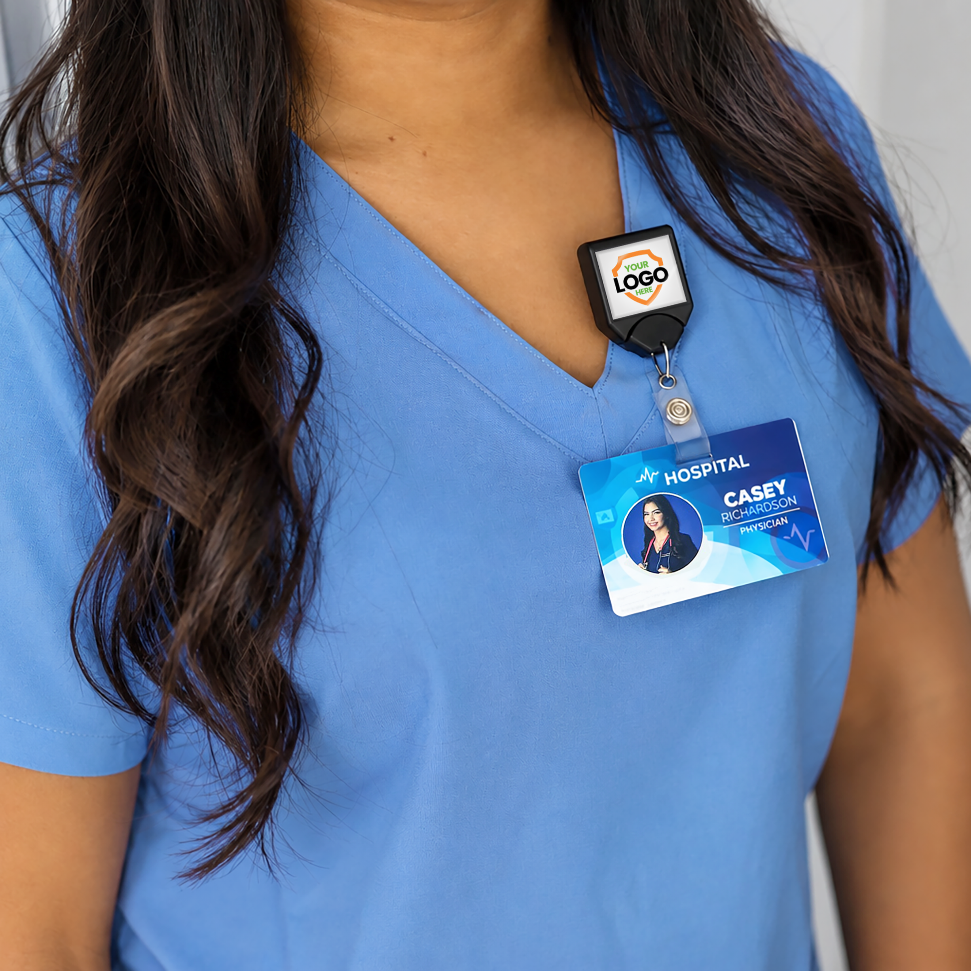 A person in blue scrubs wears a hospital ID badge attached to a Custom Printed B-REEL Retractable Badge Reel with Saw-Toothed Swivel-clip featuring their company logo; their long dark hair is visible, but their face is not shown.