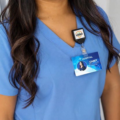 A person in blue scrubs wears a hospital ID badge attached to a Custom Printed B-REEL Retractable Badge Reel with Saw-Toothed Swivel-clip featuring their company logo; their long dark hair is visible, but their face is not shown.