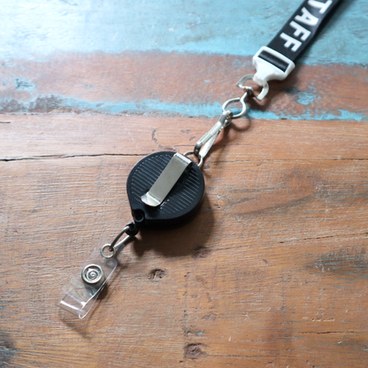 A sleek Badge Reel with Lanyard Attachment and Belt Clip (P/N 2124-302X) featuring a retractable staff holder is seamlessly joined to a lanyard, resting on a wooden surface. Sold by Specialist ID.