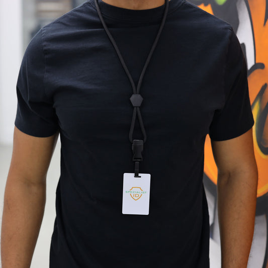 A person is wearing a plain black t-shirt and a Metal Detector Friendly Heavy Duty Lanyard with Diamond Slider, Breakaway Clasp & Quick Release Plastic Hook - MRI Safe (2135-45XX), which holds a white ID badge; they are standing indoors with part of a colorful mural visible in the background.