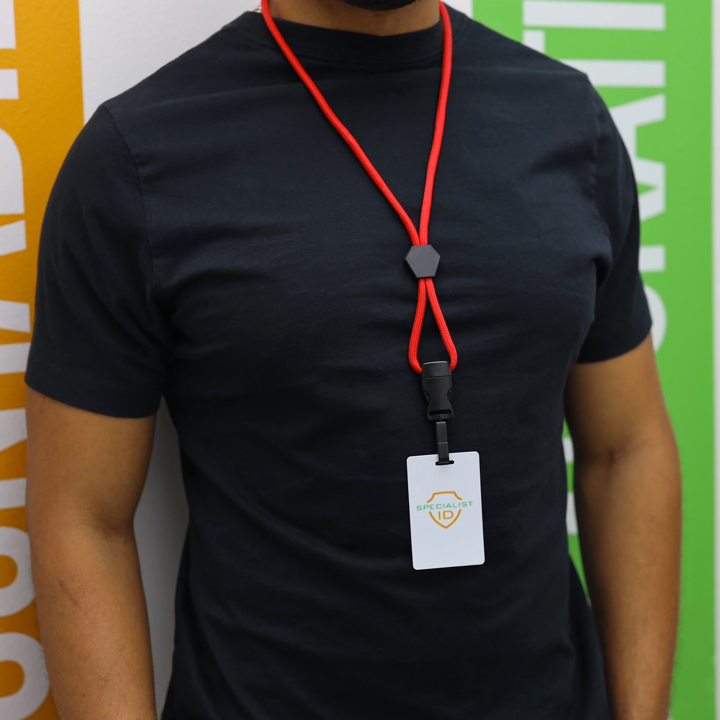 A person wearing a plain black t-shirt and a Metal Detector Friendly Heavy Duty Lanyard with Diamond Slider, Breakaway Clasp & Quick Release Plastic Hook (MRI Safe, 2135-45XX) displaying an ID badge, stands in front of colorful vertical signs.