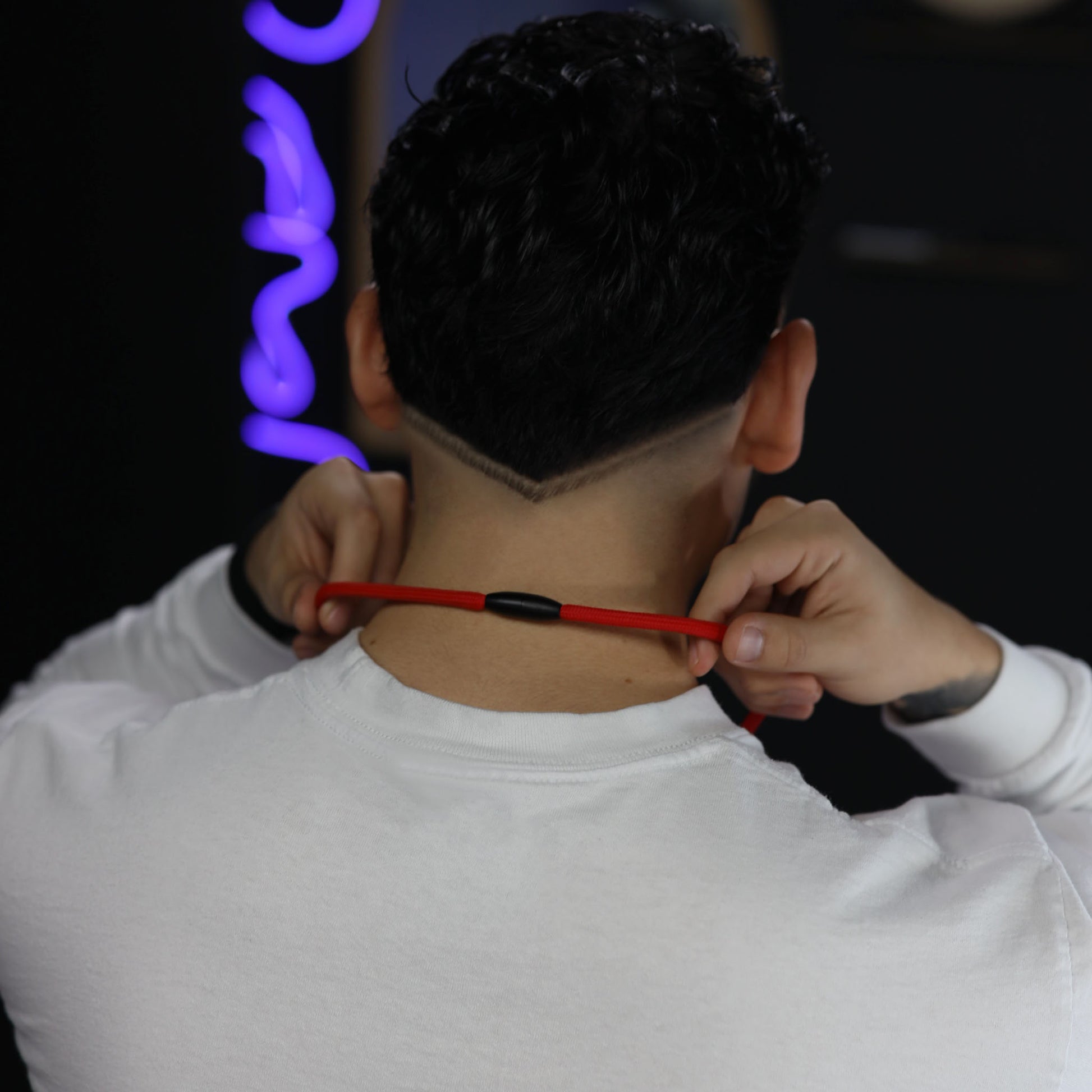 A person with a precise V-shaped nape haircut, wearing a white shirt, adjusts a Metal Detector Friendly Heavy Duty Lanyard with Diamond Slider, Breakaway Clasp & Quick Release Plastic Hook – MRI Safe (2135-45XX) around their neck. The background is dark, accented by blurred purple and blue lights.