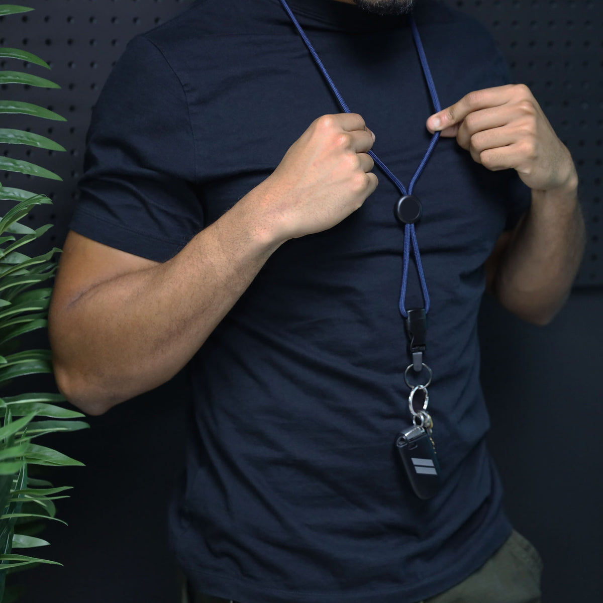 A person in a black shirt wears the 1/4″ Breakaway Round Lanyard with Detachable Split Ring (2135-461X) around their neck, with a key fob attached to the split ring. A green plant appears on the left side.