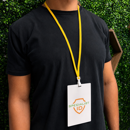 Person wearing a black t-shirt and a yellow Breakaway Lanyard with No Twist Plastic ID Clip (2137-474X), MRI and metal detector friendly, with a white Specialist ID badge, standing before a green leafy wall.