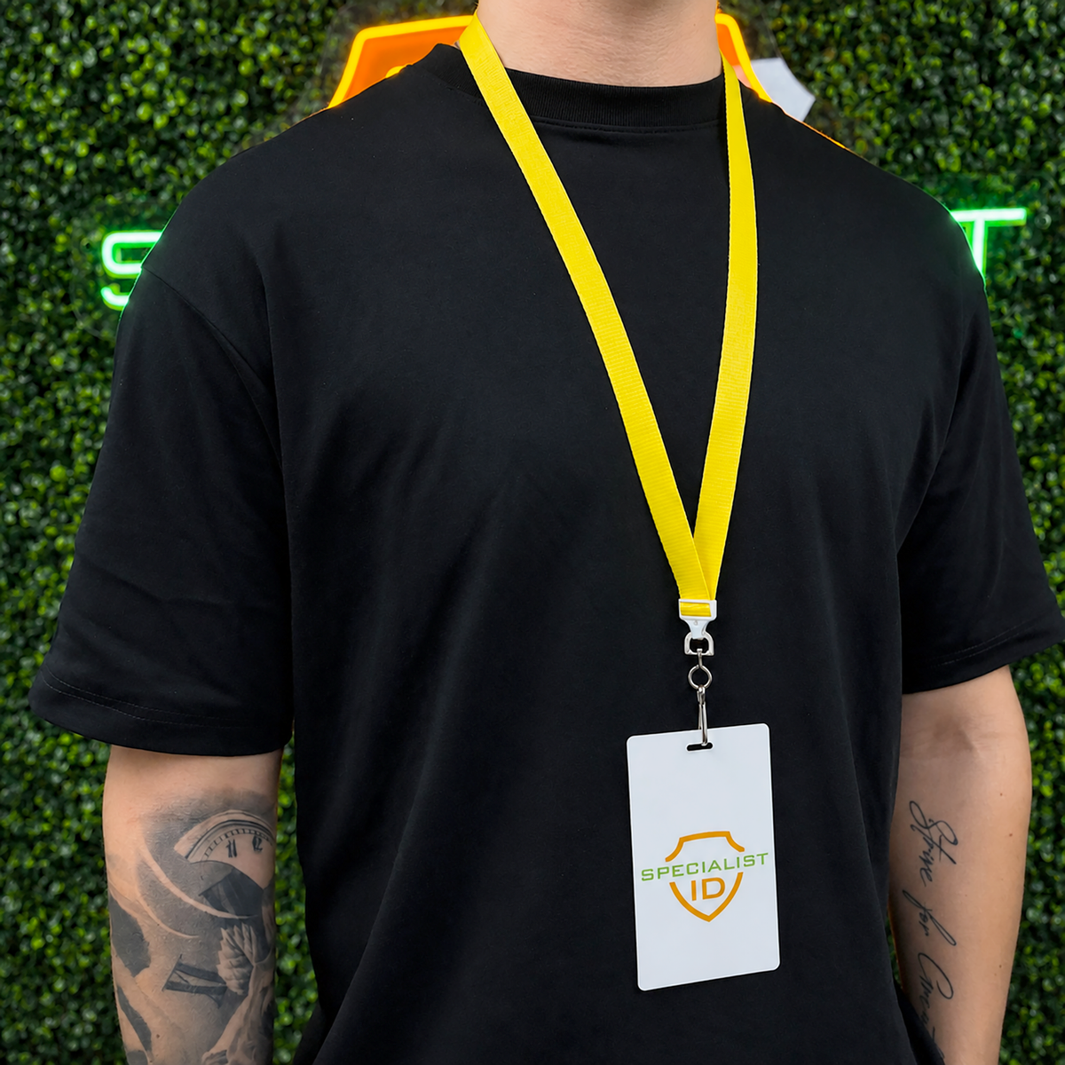 A person in a black t-shirt wears a yellow Flat Wide Breakaway Lanyard with Swivel Hook - 5/8 Inch Width (2138-500X) and Specialist ID badge, standing before green leafy decor and a neon sign.