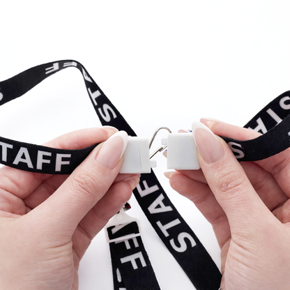 Two hands attach the clip of the black STAFF Lanyard with Standard Swivel Hook 2138-5210, featuring "STAFF" printed in white letters, against a white background.
