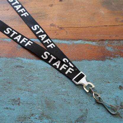 STAFF Lanyard with Standard Swivel Hook 2138-5210, displaying "STAFF" in white letters on a black background, laid out flat on a distressed wooden surface, featuring a durable swivel hook attachment.